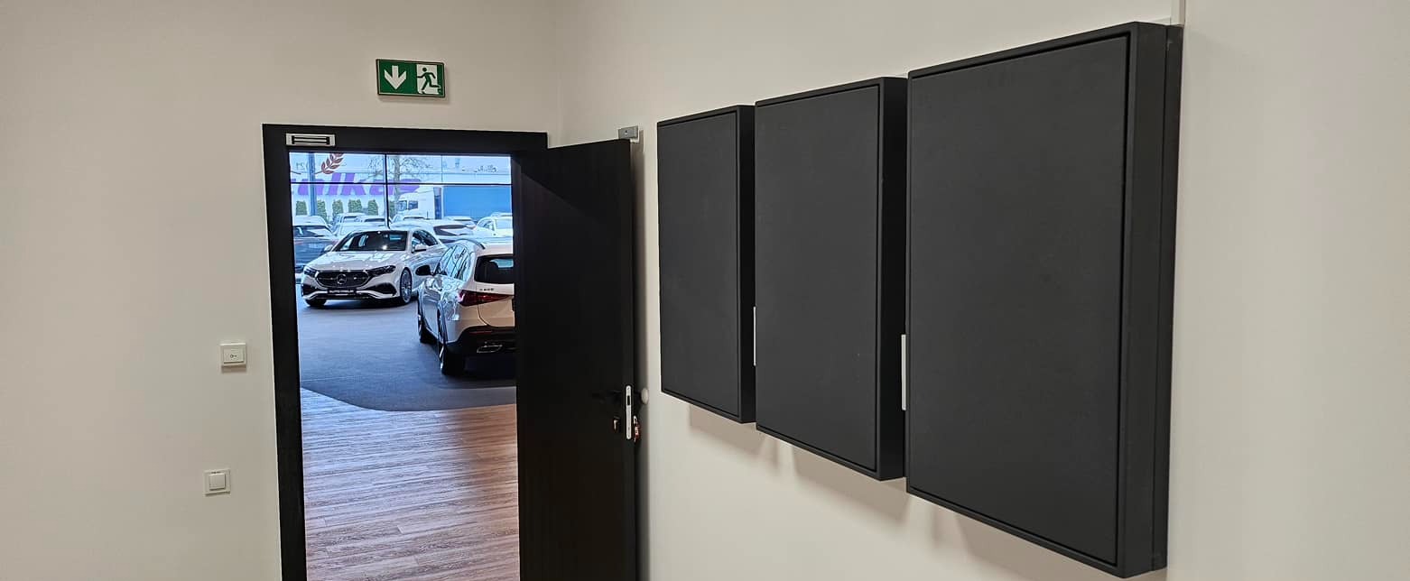 Trzy czarne szafki wiszące na białej ścianie, widoczne przy wejściu do salonu samochodowego z zaparkowanymi autami.