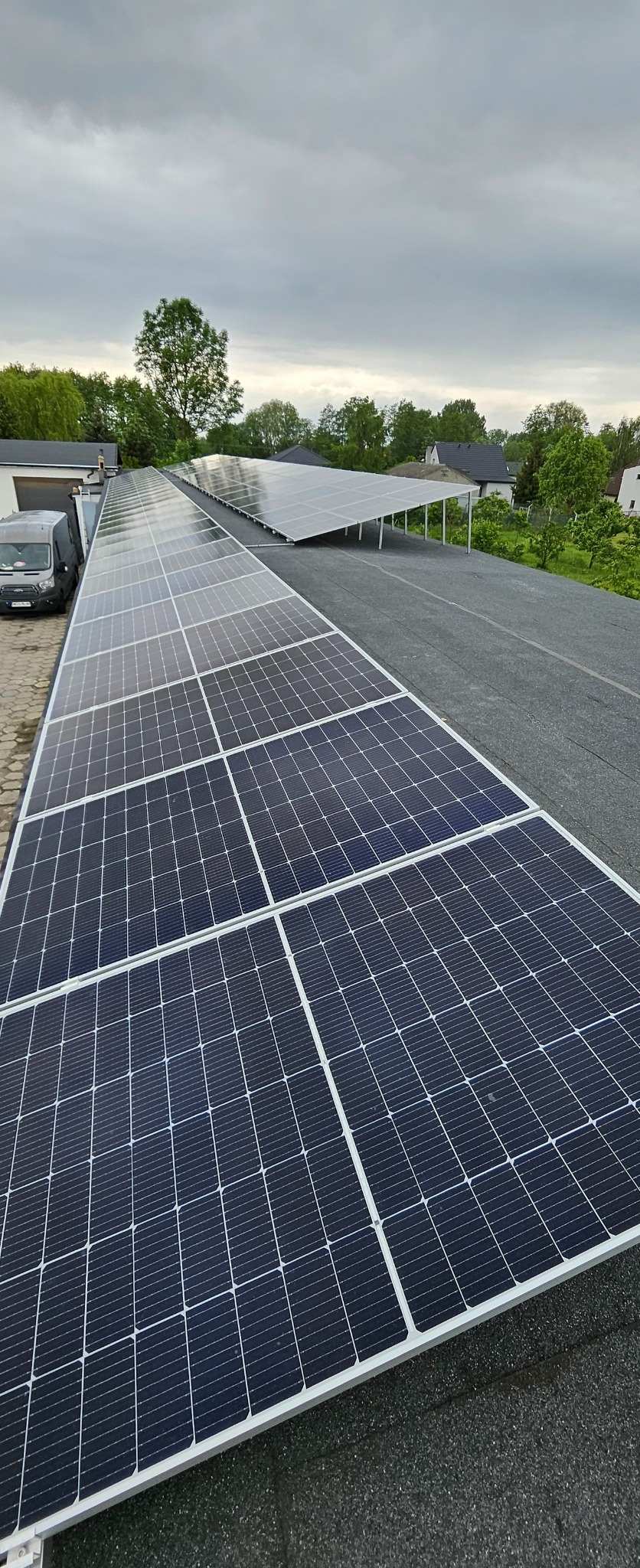 Rząd paneli fotowoltaicznych na płaskim dachu z widocznym samochodem dostawczym w tle oraz konstrukcją wsporczą dla kolejnych paneli.