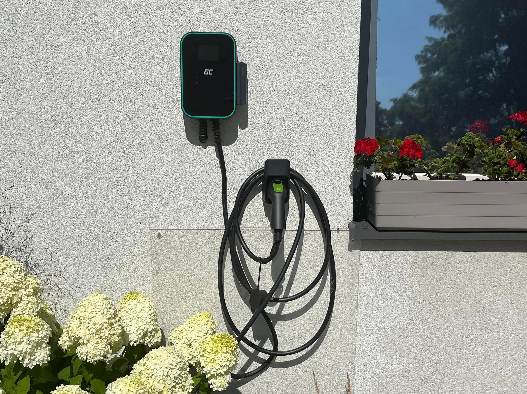 Na elewacji domu zamontowana stacja ładowania pojazdów elektrycznych marki GC z podpiętym kablem, obok kwitnące hortensje i skrzynka z czerwonymi pelargoniami w oknie.