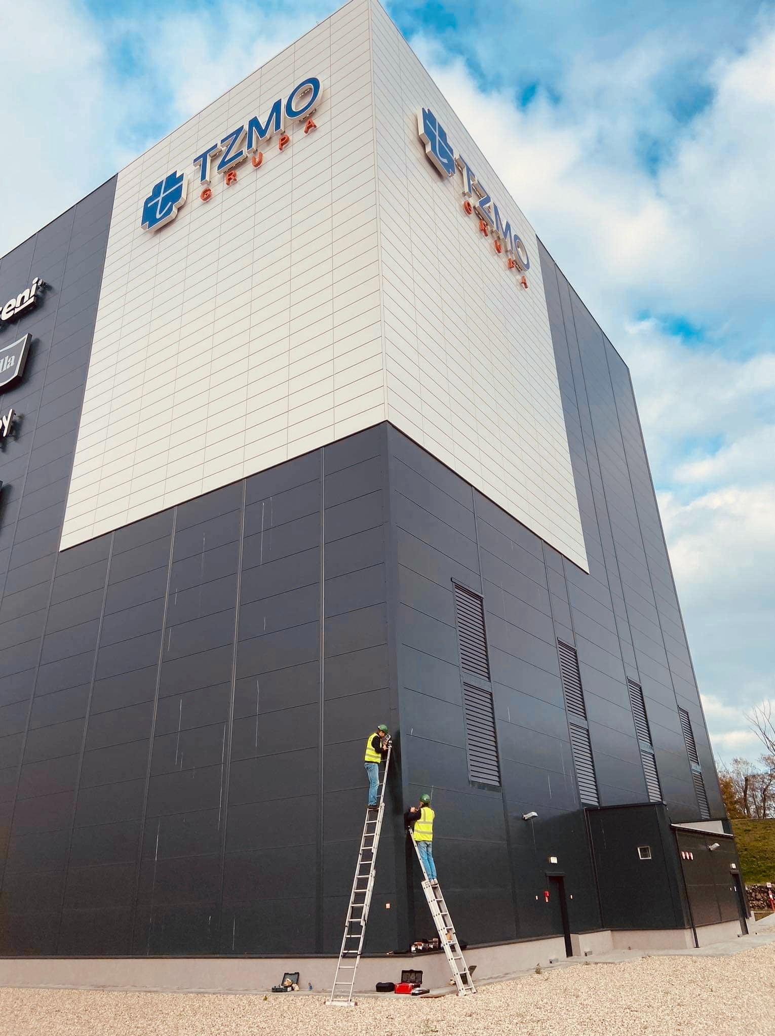 Dwóch pracowników na drabinach montuje lub naprawia elementy elewacji budynku z logo TZMO Grupa, na tle błękitnego nieba z chmurami.
