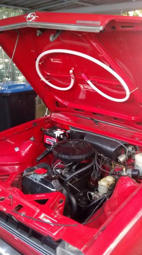 Otwarta maska czerwonego, zabytkowego samochodu z widocznym silnikiem, akumulatorem Perion i białą rurką podtrzymującą maskę.