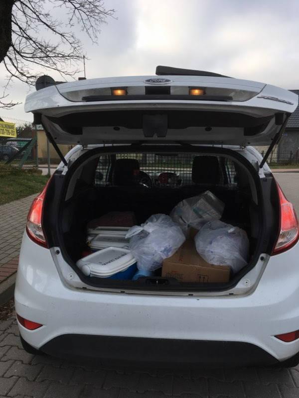 Otwarty bagażnik białego samochodu Ford Fiesta, wypełniony różnymi przedmiotami, w tym pojemnikiem chłodzącym, kartonem i workami z tworzywa sztucznego. Widoczna siatka zabezpieczająca. Ulica w tle.