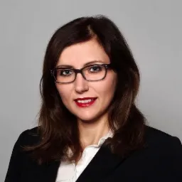 Portret kobiety w okularach i czarnym żakiecie, z czerwonymi ustami, na szarym tle.