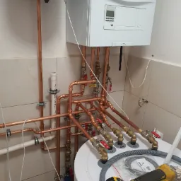 Instalacja kotła grzewczego Vaillant z miedzianymi rurami, zaworami i zbiornikiem, widoczna szlifierka kątowa DeWalt na zbiorniku.