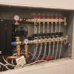USŁUGI INSTALACYJNO BUDOWLANE SEBASTIAN WÓJCIK - Rozdzielacz ogrzewania podłogowego zamontowany w obudowie, widoczne rury doprowadzające wodę, zawory z termostatami, manometry i sterownik elektroniczny ENGO, całość zainstalowana w szarej wnęce ze...
