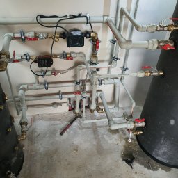 USŁUGI INSTALACYJNO BUDOWLANE SEBASTIAN WÓJCIK - Złożona instalacja hydrauliczna z rur PVC i miedzianych, z zaworami i pompą obiegową Keller, zamontowana na ścianie, z widocznymi narzędziami na podłodze, w surowym stanie wykończenia.