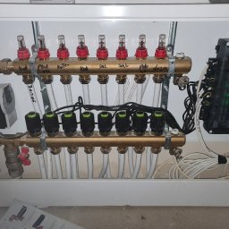 USŁUGI INSTALACYJNO BUDOWLANE SEBASTIAN WÓJCIK - Rozdzielacz ogrzewania podłogowego z widocznymi przepływomierzami, zaworami termostatycznymi, pompą obiegową i sterownikiem elektronicznym Salus, zainstalowany w białej szafce.