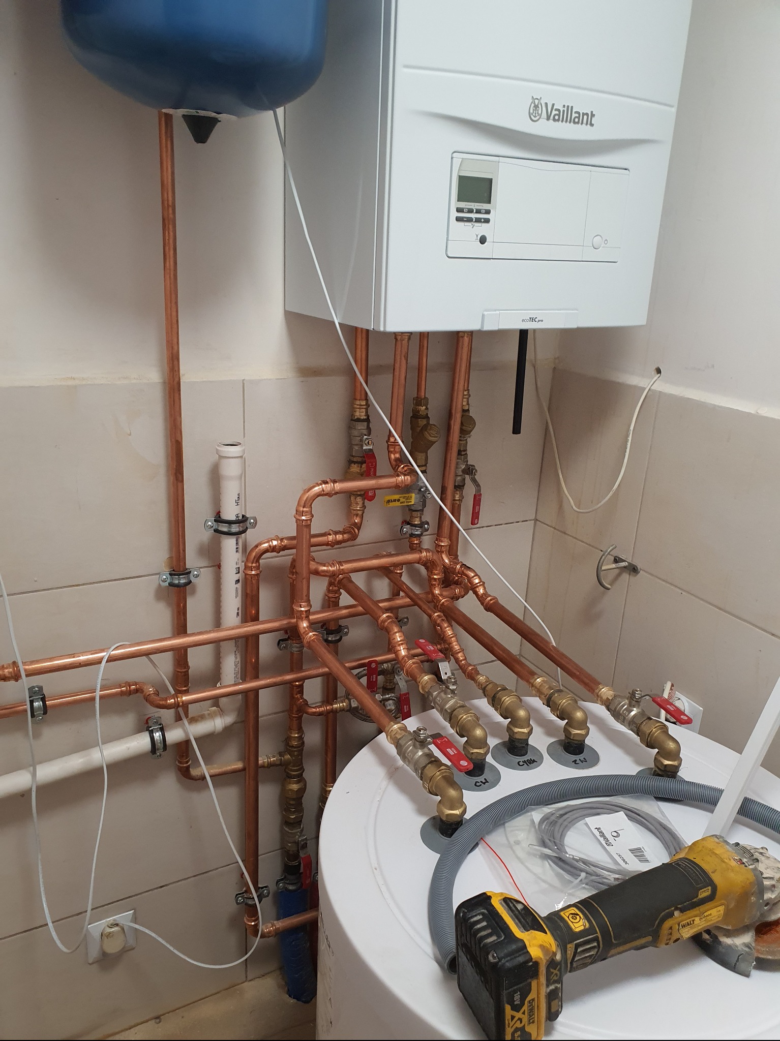 Instalacja kotła grzewczego Vaillant z miedzianymi rurami, zaworami i zbiornikiem, widoczna szlifierka kątowa DeWalt na zbiorniku.