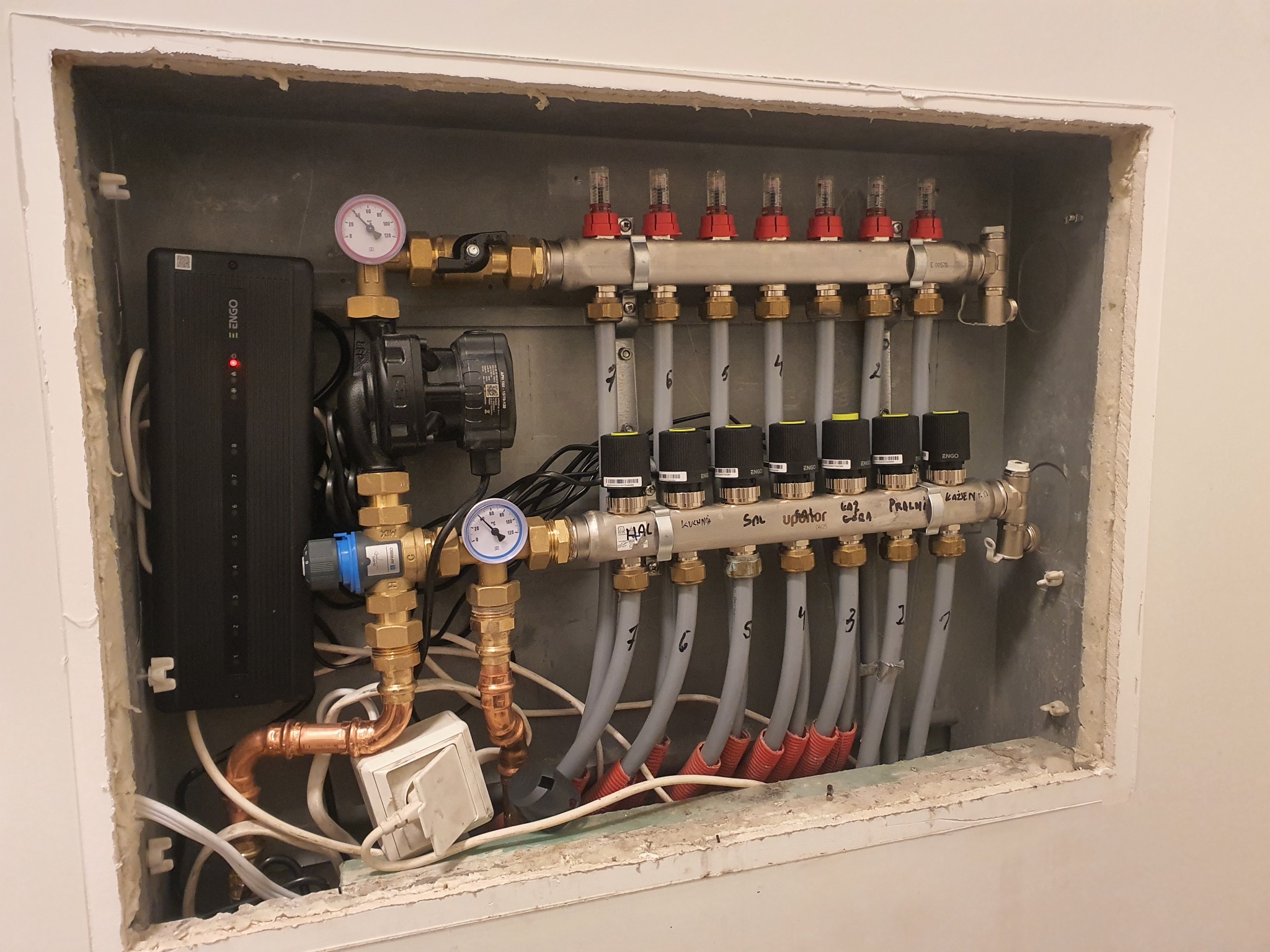 Rozdzielacz ogrzewania podłogowego zamontowany w obudowie, widoczne rury doprowadzające wodę, zawory z termostatami, manometry i sterownik elektroniczny ENGO, całość zainstalowana w szarej wnęce ze...