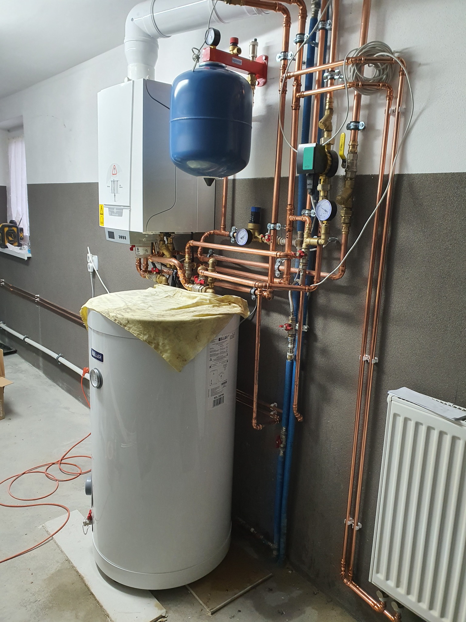 Kotłownia z nową instalacją grzewczą: piec, zasobnik ciepłej wody przykryty płachtą, miedziane rury, manometry i zawory.