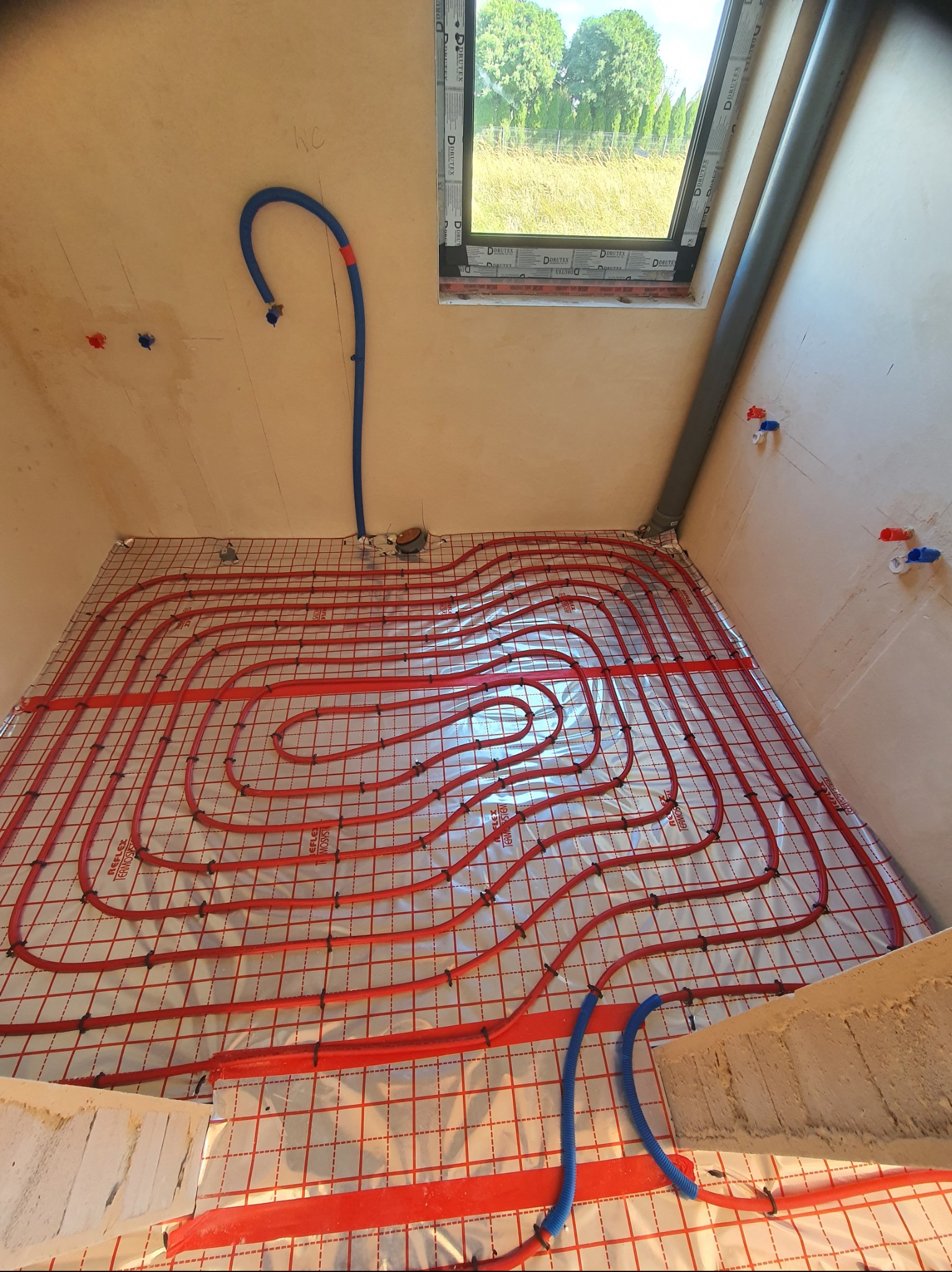 Instalacja ogrzewania podłogowego w trakcie montażu: czerwone rurki ułożone spiralnie na srebrnej folii odblaskowej z siatką, widok z góry, nieotynkowane ściany i okno dachowe w tle.