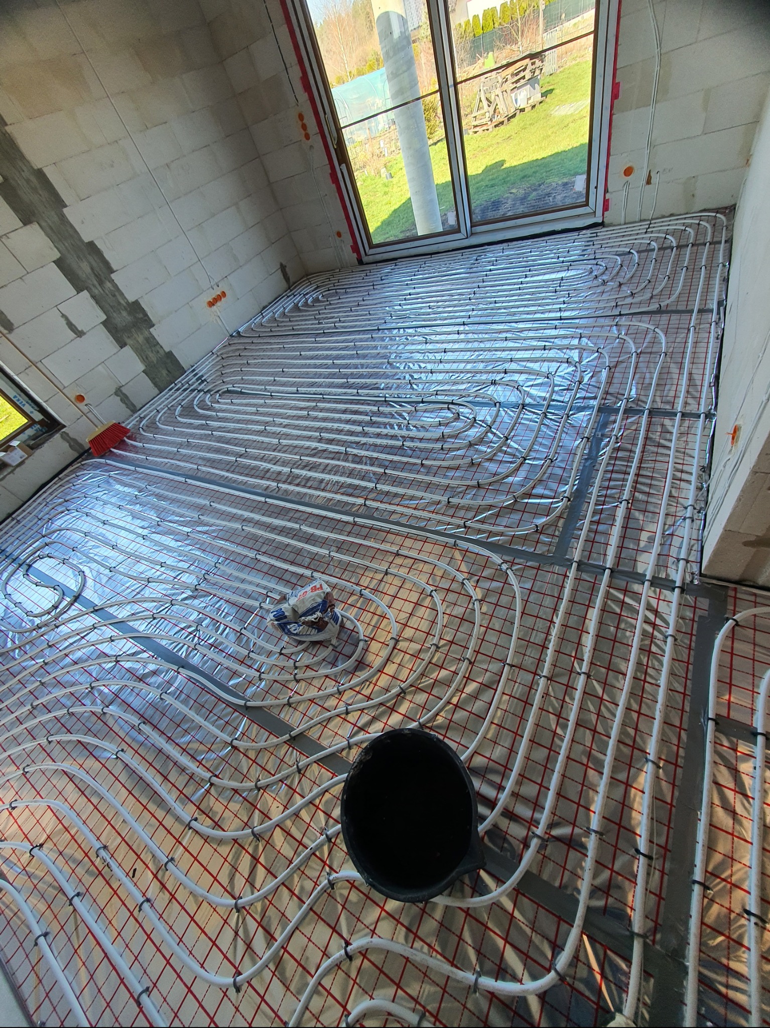 Białe rury ogrzewania podłogowego ułożone na folii z czerwoną kratką, widok na okno i surowy stan ścian z betonu komórkowego.