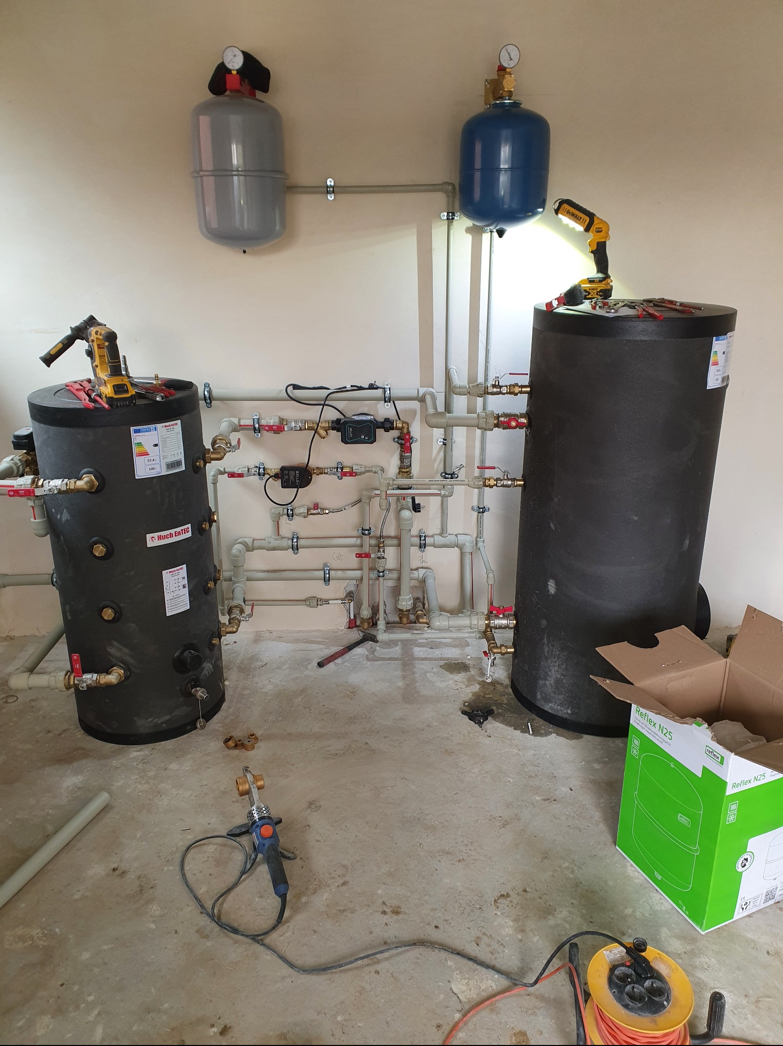 Instalacja dwóch czarnych zbiorników akumulacyjnych z podłączonymi rurami i zaworami, widoczne narzędzia hydrauliczne i skrzynka Reflex N25, manometry na ścianie.