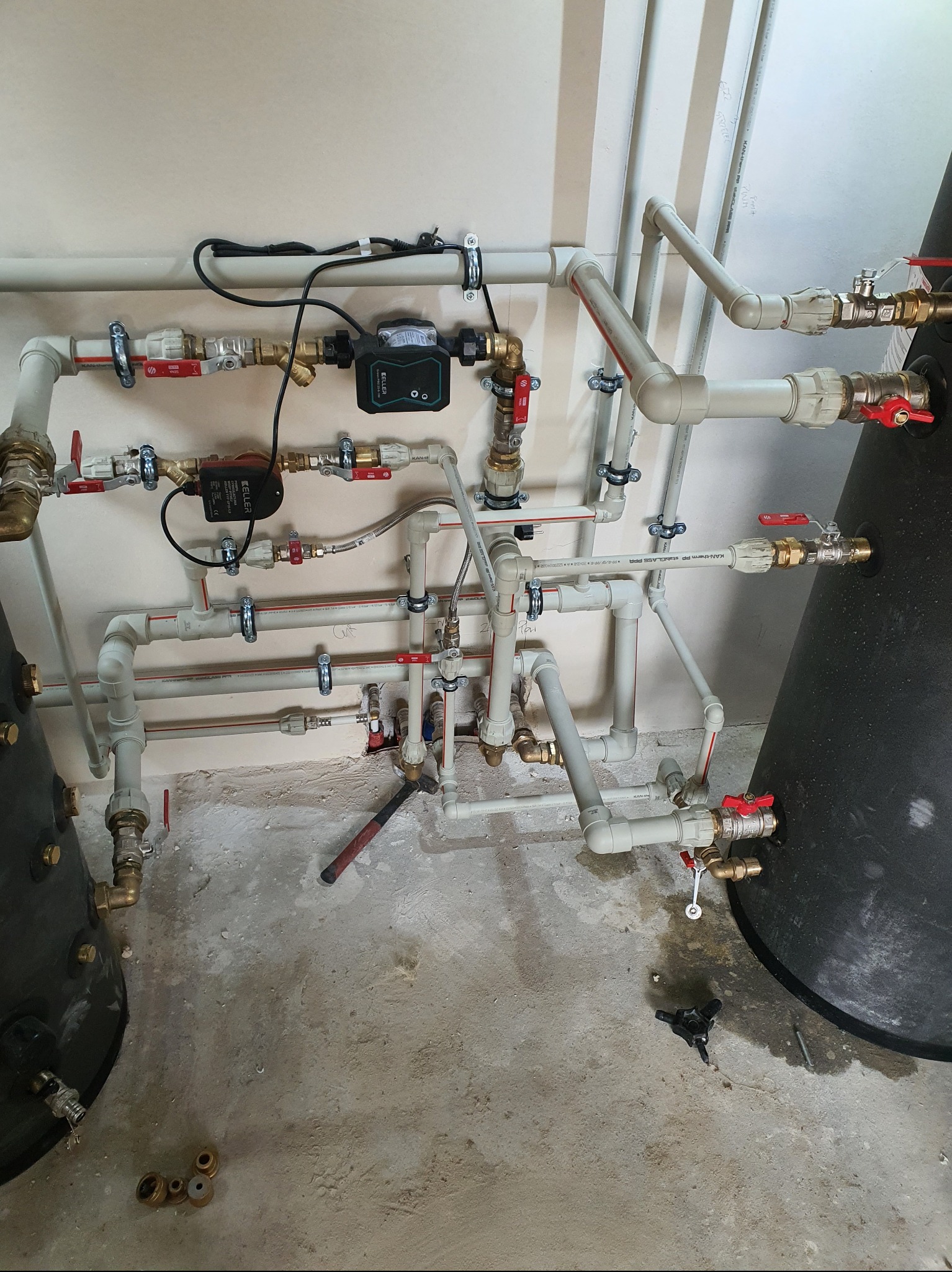 Złożona instalacja hydrauliczna z rur PVC i miedzianych, z zaworami i pompą obiegową Keller, zamontowana na ścianie, z widocznymi narzędziami na podłodze, w surowym stanie wykończenia.