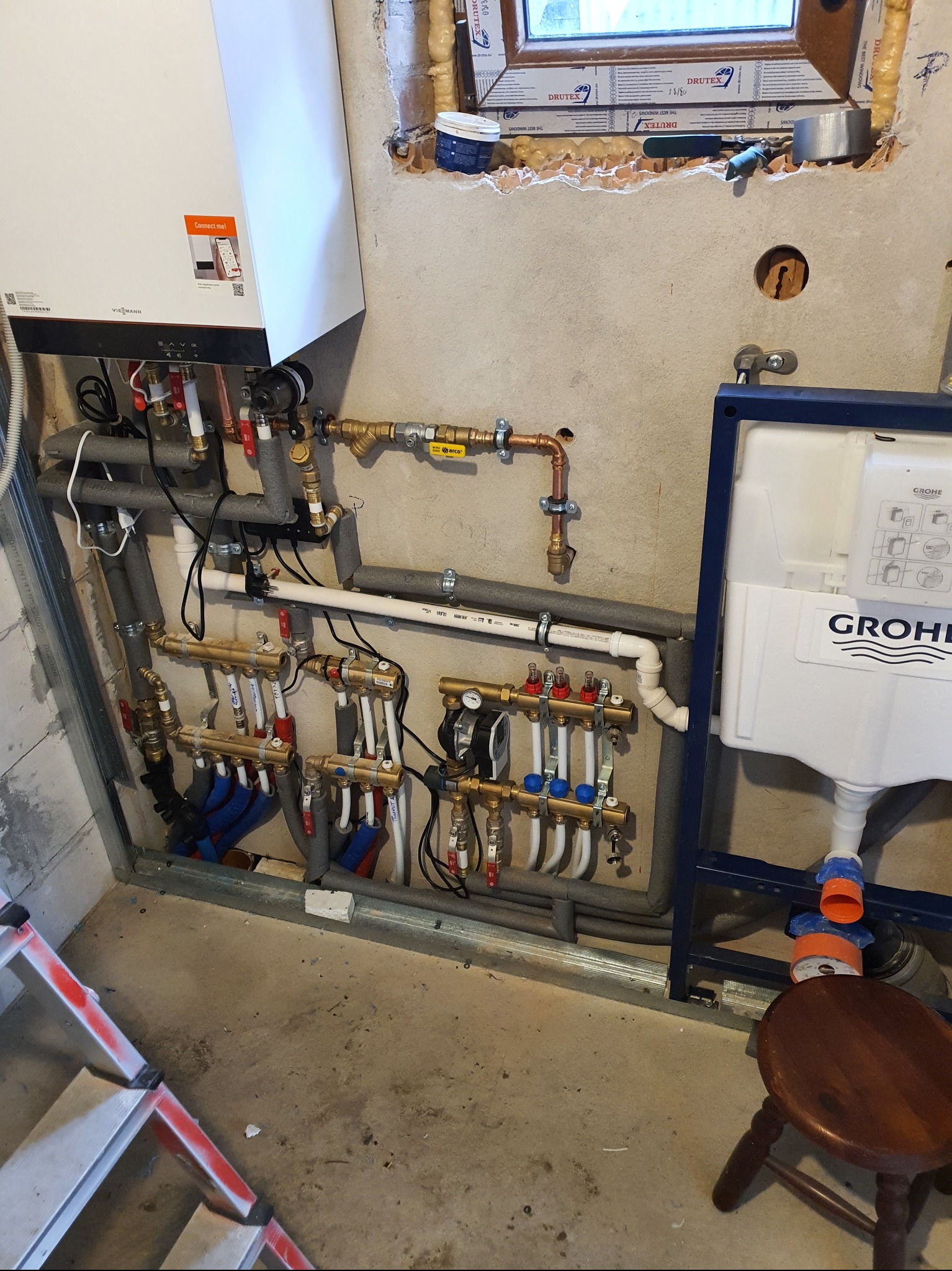 Instalacja grzewcza z kotłem Viessmann, rozdzielaczem ogrzewania podłogowego i stelażem podtynkowym Grohe, widoczne rury w izolacji, drabina i stołek w surowym pomieszczeniu.