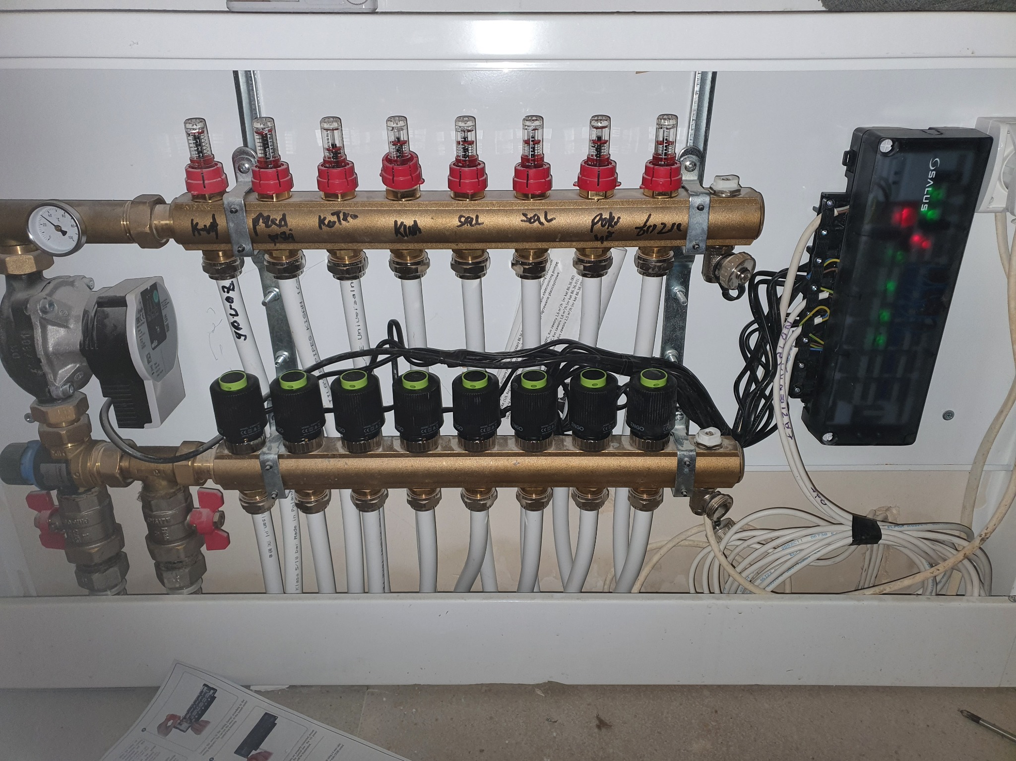 Rozdzielacz ogrzewania podłogowego z widocznymi przepływomierzami, zaworami termostatycznymi, pompą obiegową i sterownikiem elektronicznym Salus, zainstalowany w białej szafce.