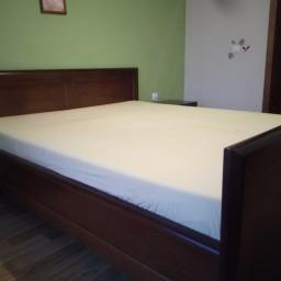 MEBLO-STYL JACEK CHACHUŁA - Klasyczne łóżko z ciemnego drewna z pościelą w jasnym odcieniu, widoczne na tle zielonej i białej ściany z drobnymi dekoracjami.