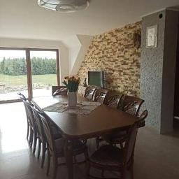 MEBLO-STYL JACEK CHACHUŁA - Duży, drewniany stół jadalny z ośmioma krzesłami w przestronnym wnętrzu z widokiem na zewnątrz, na stole wazon z tulipanami, ściana wykończona dekoracyjną cegłą.