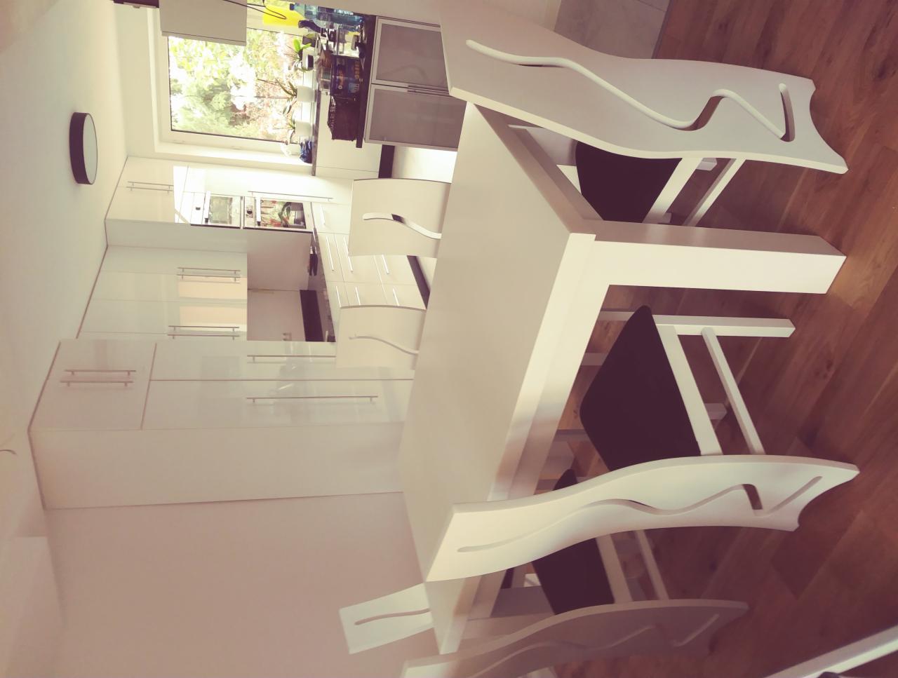 Nowoczesna biała kuchnia z minimalistycznymi szafkami, białym stołem i krzesłami z falistym oparciem, widok z boku na drewnianą podłogę.