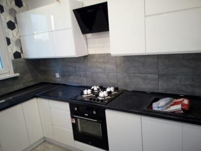 Nowoczesna kuchnia z białymi szafkami, czarnym blatem i płytą grzewczą z garnkami, szare płytki na ścianie, widok z lekkiego ukosa.