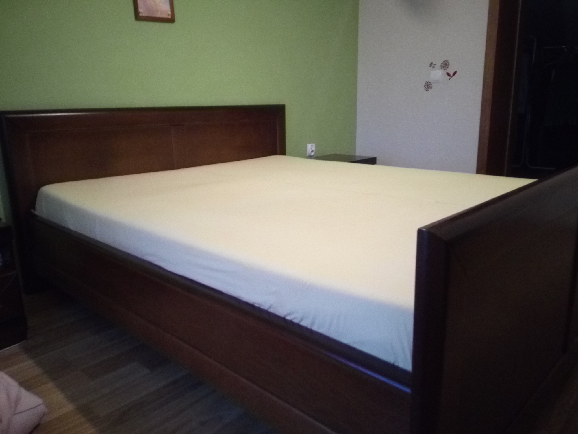 Klasyczne łóżko z ciemnego drewna z pościelą w jasnym odcieniu, widoczne na tle zielonej i białej ściany z drobnymi dekoracjami.