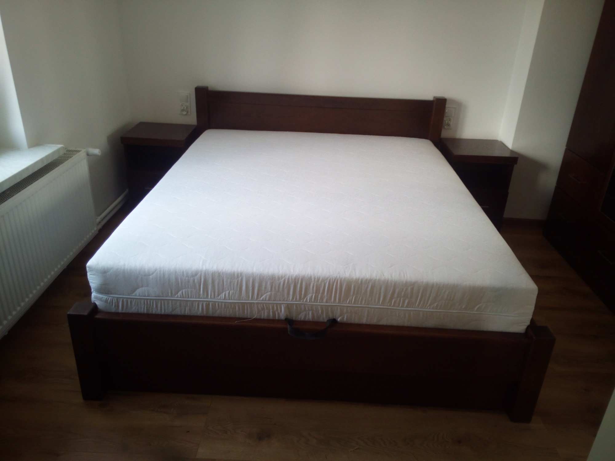 Ciemnobrązowe łóżko z drewnianą ramą i zagłówkiem, z białym materacem, szafki nocne po bokach, na drewnianej podłodze.