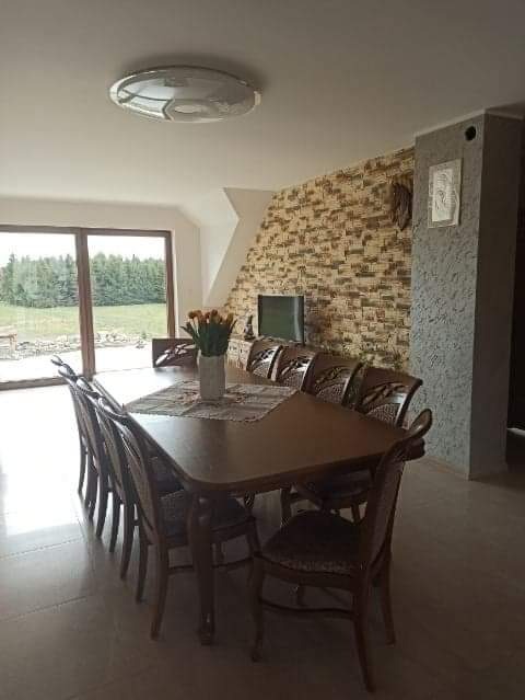 Duży, drewniany stół jadalny z ośmioma krzesłami w przestronnym wnętrzu z widokiem na zewnątrz, na stole wazon z tulipanami, ściana wykończona dekoracyjną cegłą.