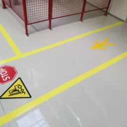 Jasnoszara podłoga w hali produkcyjnej z żółtymi liniami wyznaczającymi ciągi komunikacyjne, znak ostrzegawczy z wózkiem widłowym i znak STOP w lewym dolnym rogu oraz sylwetka pieszego w centralnej...