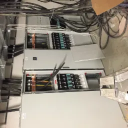 Rozdzielnica elektryczna w trakcie instalacji, widoczne kable, szyny DIN, zabezpieczenia i drabina monterska w tle, ukazująca etap prac instalacyjnych.