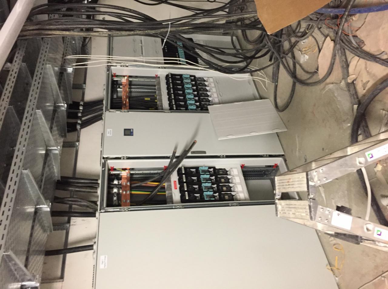 Rozdzielnica elektryczna w trakcie instalacji, widoczne kable, szyny DIN, zabezpieczenia i drabina monterska w tle, ukazująca etap prac instalacyjnych.