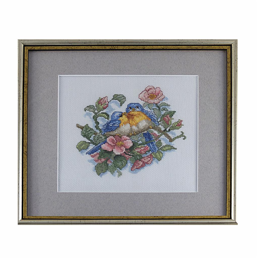 Oprawiona w ramkę haftowana kanwa przedstawiająca parę błękitnych ptaków siedzących na gałęzi z różowymi kwiatami, widoczna faktura płótna.