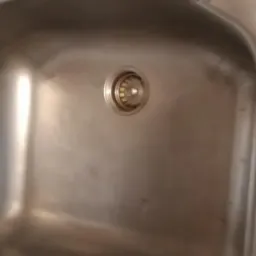 Błyszczący, stalowy zlew kuchenny z widocznym odpływem, otoczony fragmentami płytek i blatu.