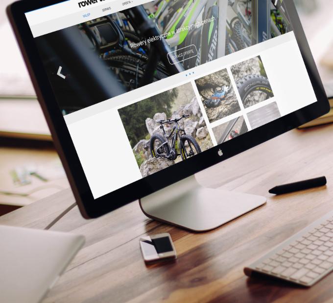Strona internetowa firmy z Bydgoszczy prezentująca rowery elektryczne na ekranie komputera iMac stojącego na biurku z klawiaturą, długopisem i smartfonem.
