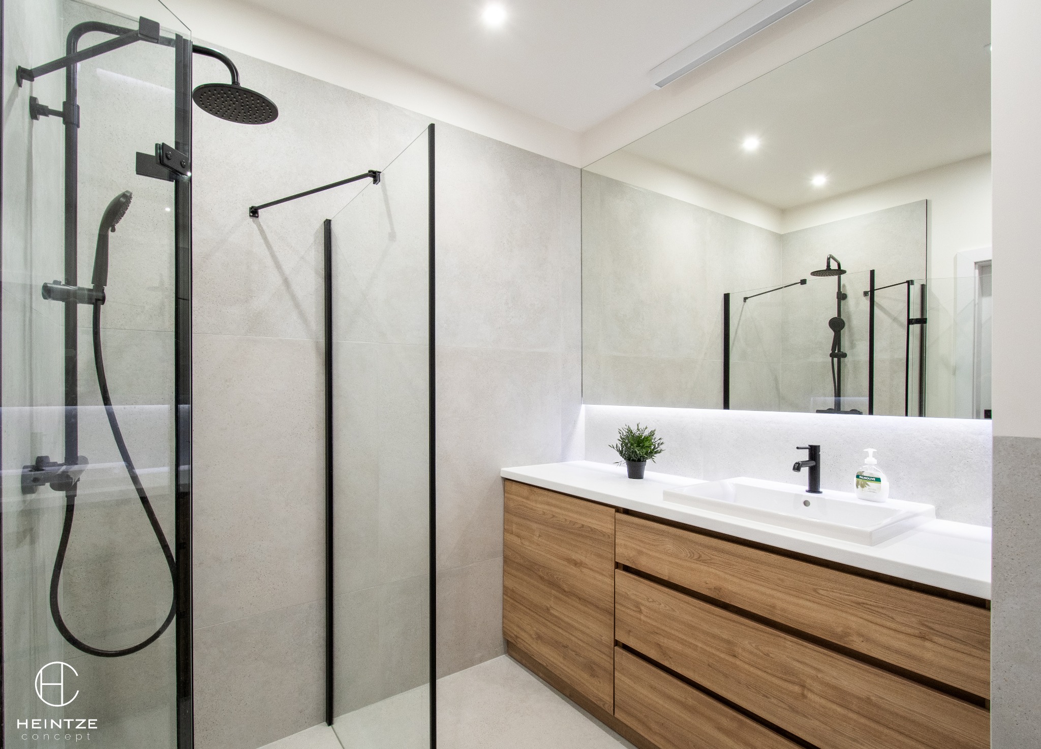 Nowoczesna łazienka z kabiną prysznicową typu walk-in z czarnymi profilami, drewnianą szafką pod umywalką z białym blatem, dużym lustrem i oświetleniem LED.