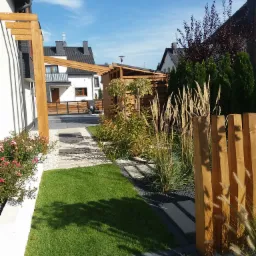 Nowoczesny ogród przydomowy z drewnianą pergolą, ścieżką z płyt betonowych, ozdobnymi trawami i białym żwirem.