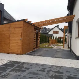 Drewniana wiata na drewno połączona z pergolą, przylegająca do domu, z widocznym ułożonym drewnem, na zewnątrz kostka brukowa i kamień ozdobny.