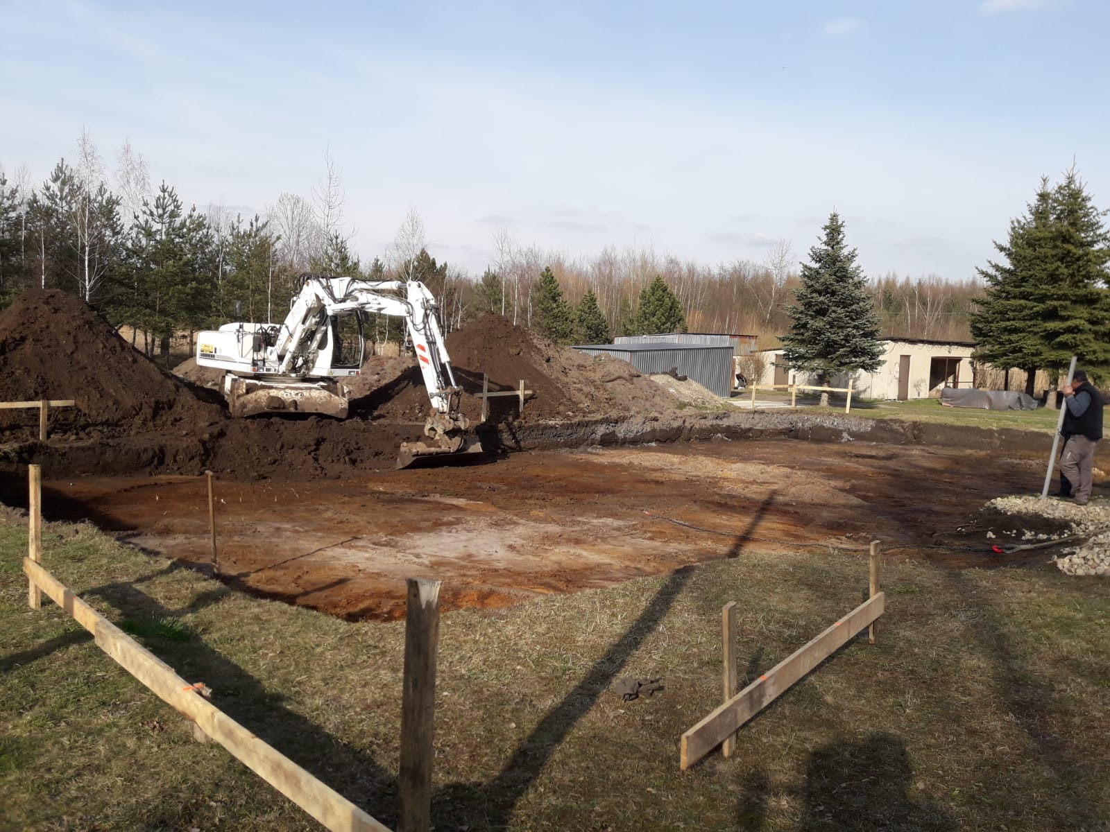 Biała koparka na placu budowy, wykop pod fundamenty zaznaczony drewnianymi słupkami, pryzmy ziemi w tle, pracownik z łatą niwelacyjną po prawej.