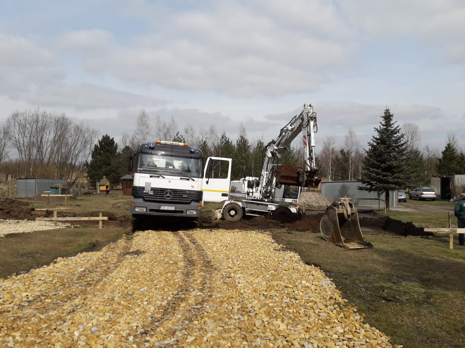 Ciężarówka Mercedes-Benz 3340 i biała koparka kołowa podczas prac ziemnych na grząskim terenie, widoczne ślady opon na żwirowej drodze.