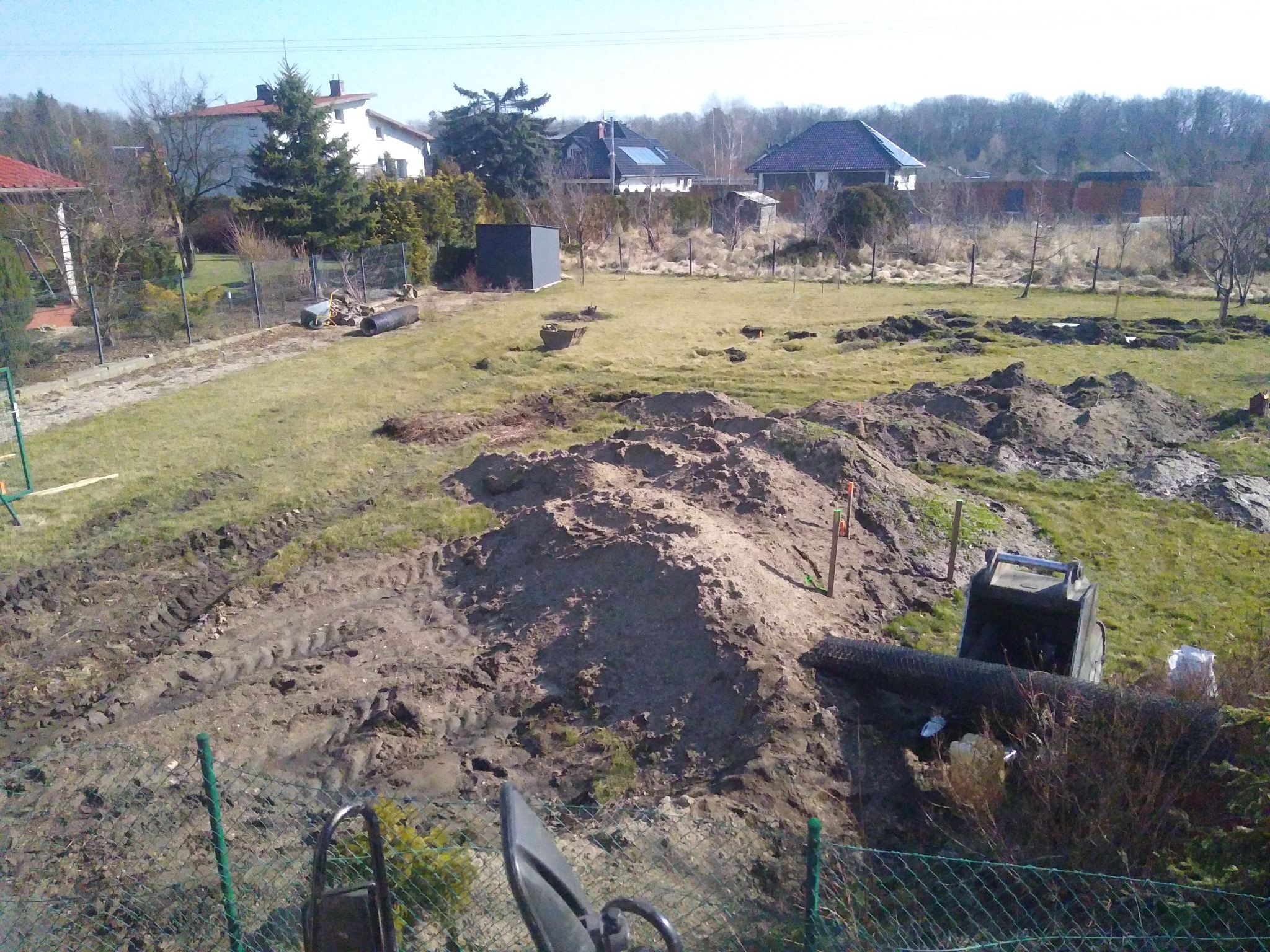 Teren po częściowym wyrównaniu z widocznymi śladami opon, niewielkim sprzętem budowlanym i stertami ziemi, ogrodzony siatką, w tle zabudowa jednorodzinna.