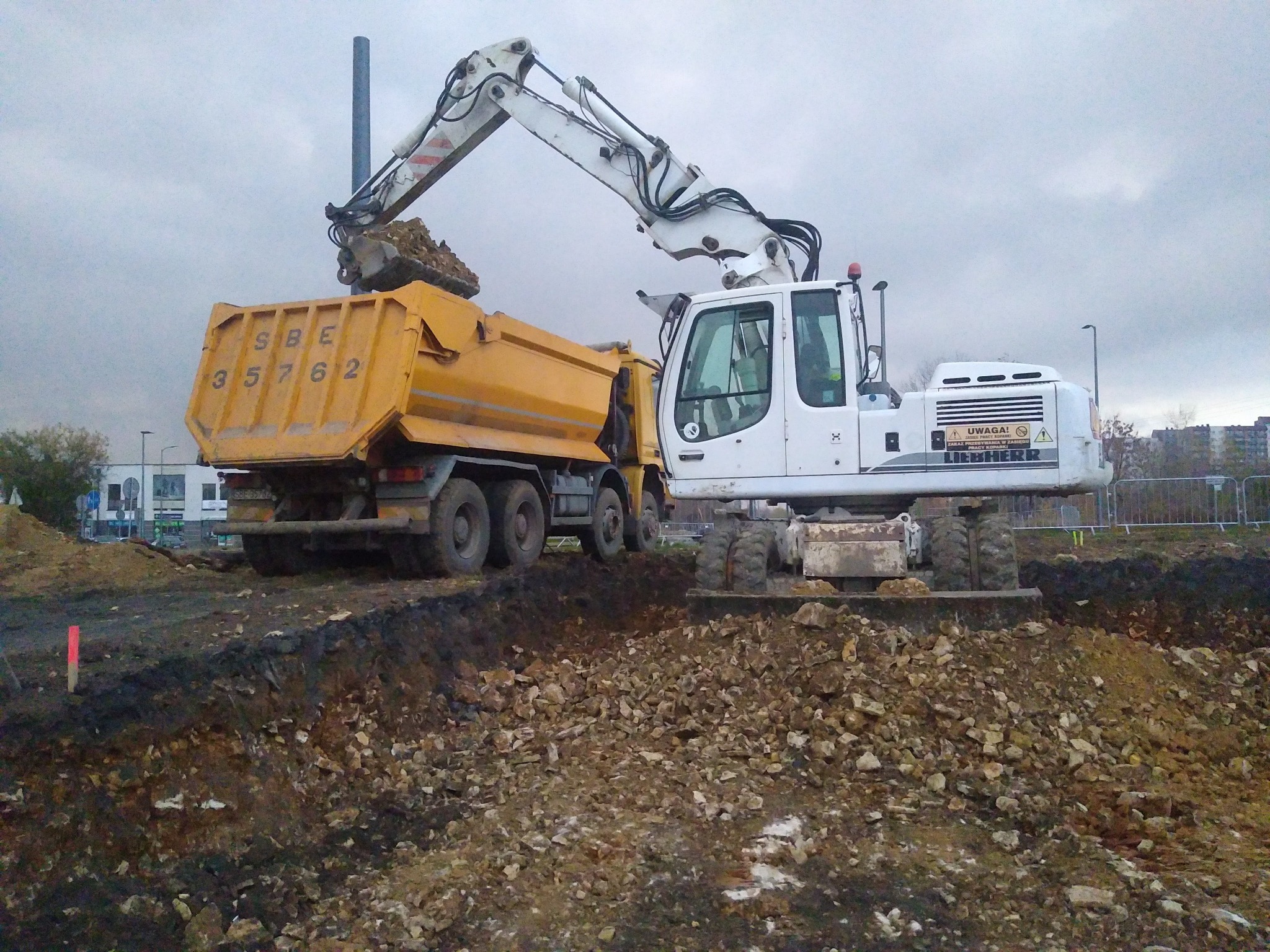 Biała koparka kołowa Liebherr ładuje urobek do żółtej ciężarówki SBE 35762 na placu budowy, widoczne świeże wykopy i sterta ziemi.