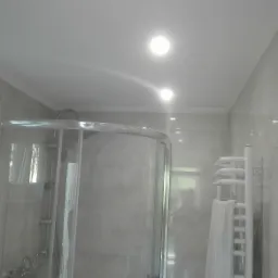 Jasna łazienka z kabiną prysznicową, grzejnikiem drabinkowym i trzema punktami oświetleniowymi w suficie. Widoczne szare płytki i białe wykończenia.