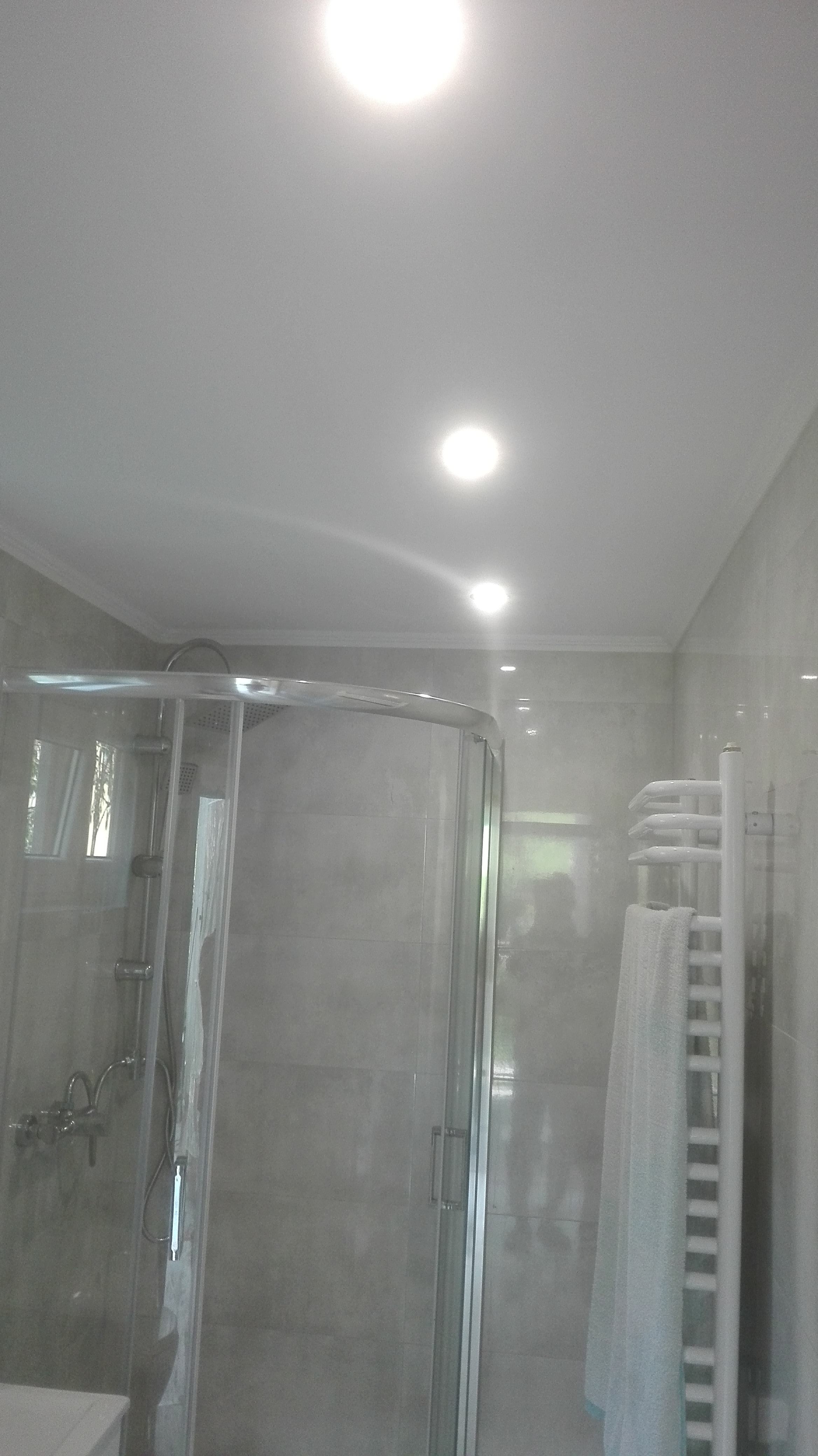 Jasna łazienka z kabiną prysznicową, grzejnikiem drabinkowym i trzema punktami oświetleniowymi w suficie. Widoczne szare płytki i białe wykończenia.