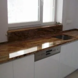 Kuchnia z blatem z polerowanego granitu w odcieniach brązu i beżu, dopasowanym do parapetu, zintegrowana ze zlewem i białą zabudową meblową.