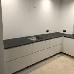 Montaż blatu kuchennego z ciemnego kamienia na białych szafkach, widoczny otwór na zlew, surowy stan wykończenia wnętrza, przygotowanie do instalacji.