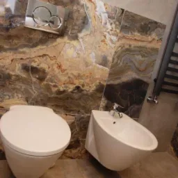 Nowoczesna łazienka w Warszawie z podwieszaną toaletą, bidetem i grzejnikiem drabinkowym na tle ściany wyłożonej marmurem o ciepłych barwach.