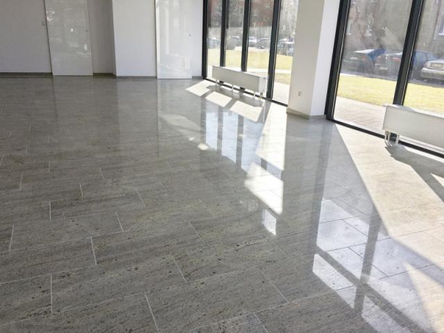 Polerowana podłoga z szarego kamienia naturalnego ułożona w prostokątną mozaikę, odbijająca światło słoneczne wpadające przez duże okna.