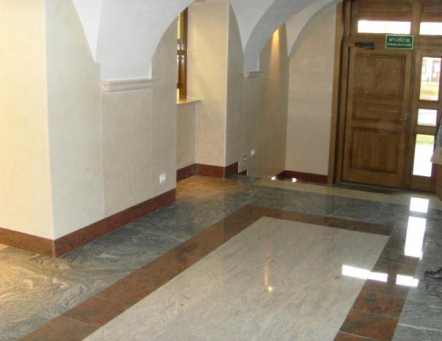 Wnętrze budynku z marmurową podłogą w odcieniach szarości i beżu, z bordiurą, kremowe ściany z łukowatymi przejściami i drewniane drzwi z zielonym znakiem ewakuacyjnym.