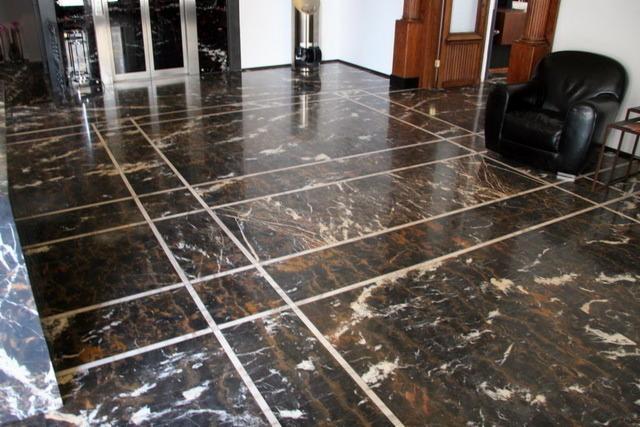 Błyszcząca posadzka z ciemnego marmuru z białymi żyłami, ułożona w kwadraty oddzielone jasnymi listwami, w tle drzwi wejściowe i skórzany fotel.