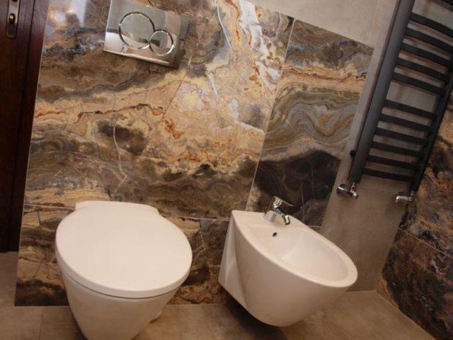 Nowoczesna łazienka w Warszawie z podwieszaną toaletą, bidetem i grzejnikiem drabinkowym na tle ściany wyłożonej marmurem o ciepłych barwach.