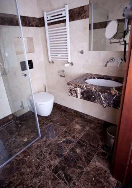 Nowoczesna łazienka z kabiną prysznicową ze szkła, wiszącą toaletą, grzejnikiem drabinkowym i umywalką nablatową z ciemnego marmuru, na podłodze brązowe płytki imitujące marmur.